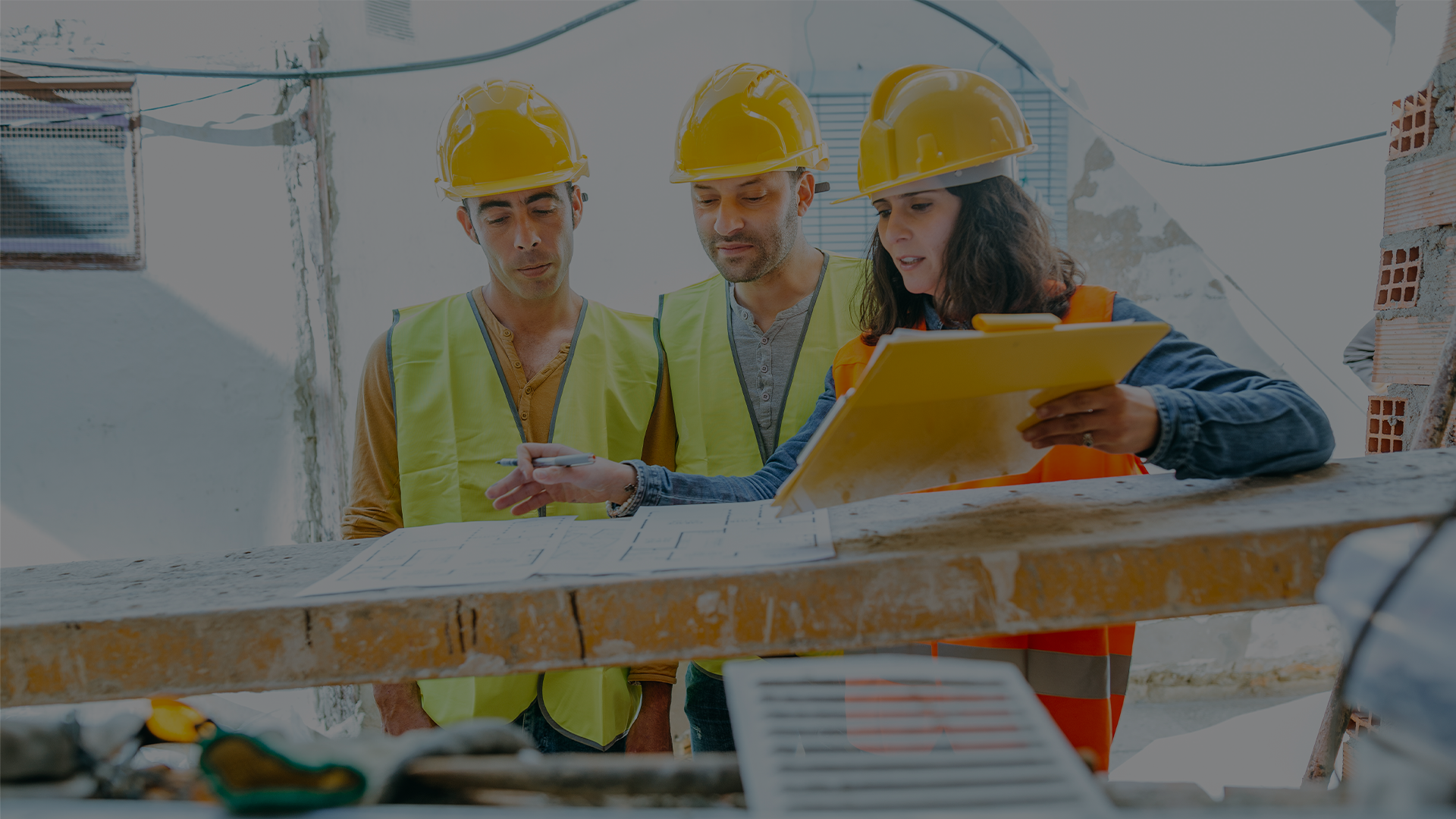 Construction safety manager reviewing a pre-task plan on a clipboard with two workers in hard hats and high-visibility vests on an active jobsite