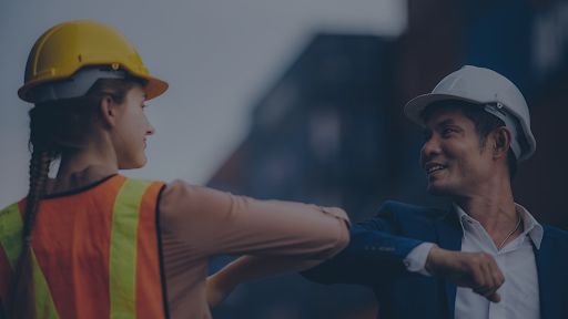 A female construction safety worker in a yellow hard hat and high-visibility vest greets a male project executive in a white hard hat with an elbow bump on an enterprise jobsite, illustrating the field-to-office connection that powers intelligent workflows and Safety Intelligence at top General Contractors.