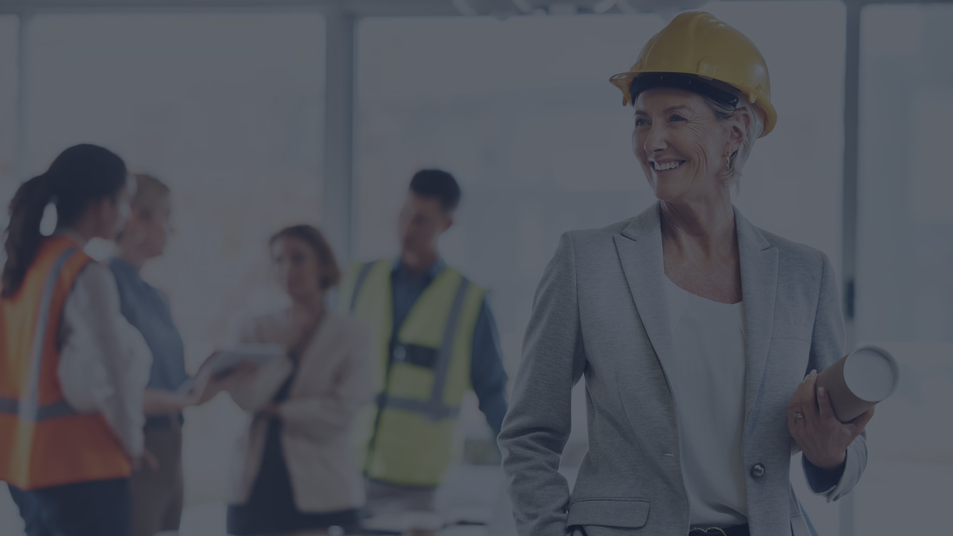 A smiling female construction executive in a yellow hard hat and blazer holds blueprints, while her team stands in the blurred background.