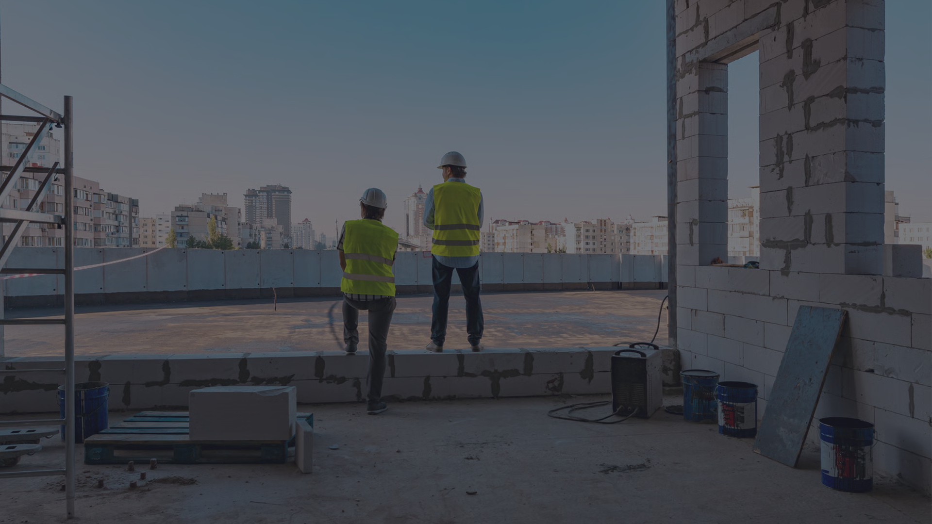 Two construction workers in hard hats and high-visibility yellow safety vests stand on a partially constructed concrete and cinderblock building, surveying the jobsite at dawn with a city skyline in the background.