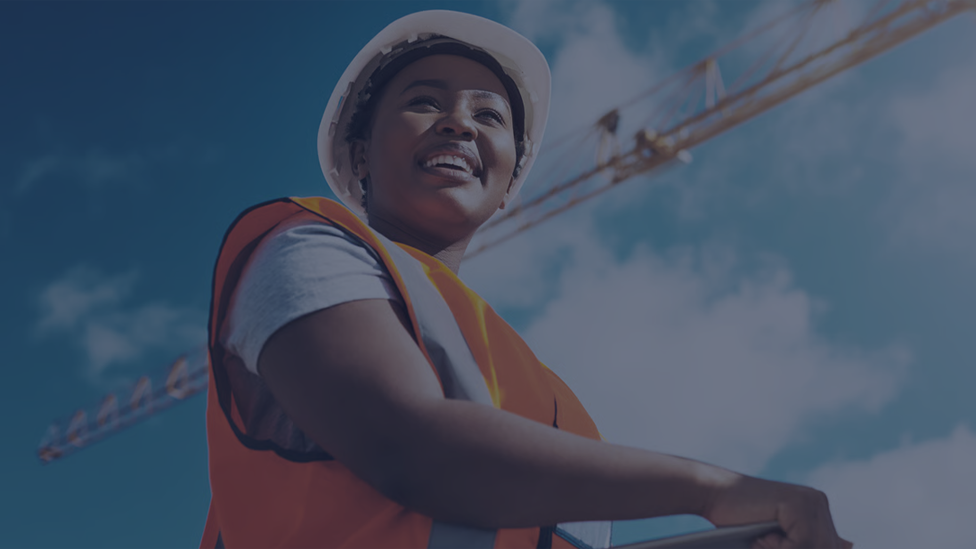 Smiling construction worker in hard hat and hi-vis vest on a jobsite with tower cranes against a blue sky, representing predictive safety management